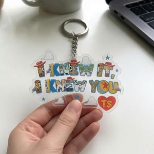 May include: A clear acrylic keyring featuring the text I KNEW IT, I KNEW YOU in a multi-coloured font with patterns inspired by cowboy and space ranger motifs. The design includes small blue stars, white cloud outlines, and a red heart with TS.