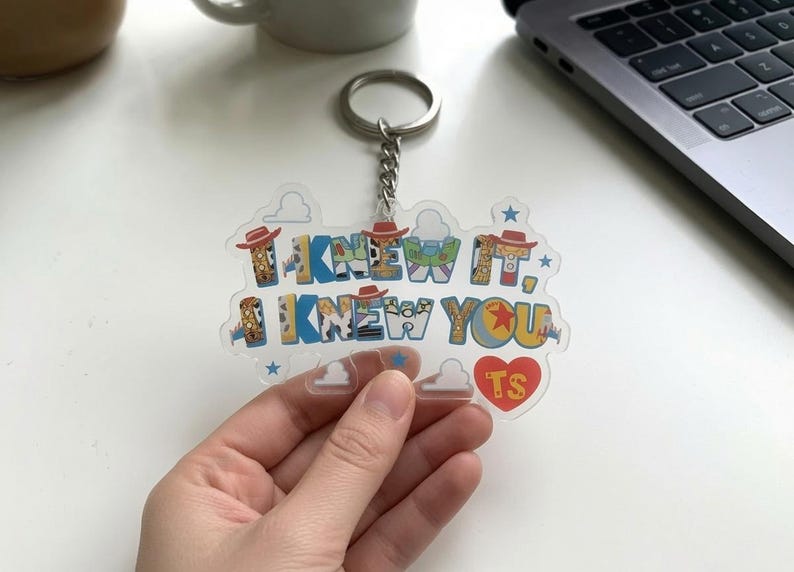 May include: A clear acrylic keyring featuring the text I KNEW IT, I KNEW YOU in a multi-coloured font with patterns inspired by cowboy and space ranger motifs. The design includes small blue stars, white cloud outlines, and a red heart with TS.