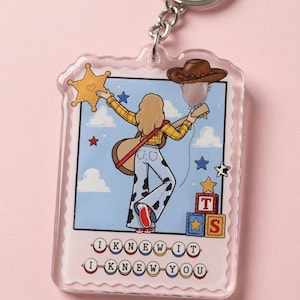 May include: A clear acrylic keychain featuring a rectangular stamp-shaped design. The illustration shows a person with blonde hair wearing a yellow plaid shirt and cow-print pants, holding a guitar. Text reads I KNEW IT I KNEW YOU.