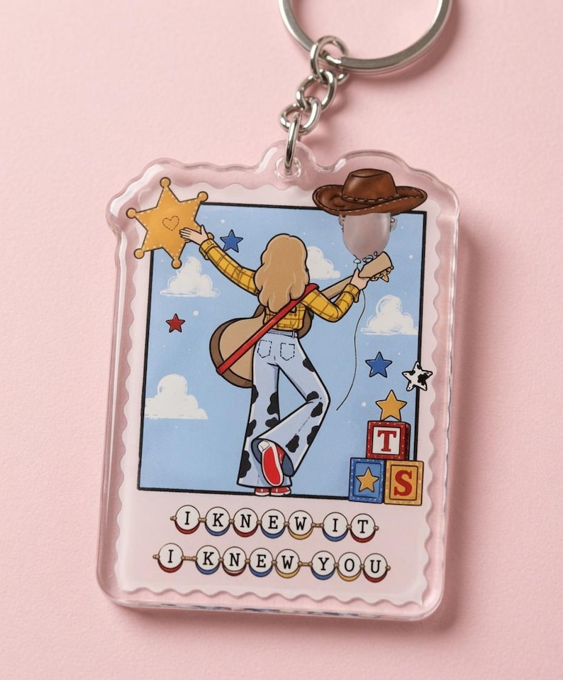May include: A clear acrylic keychain featuring a rectangular stamp-shaped design. The illustration shows a person with blonde hair wearing a yellow plaid shirt and cow-print pants, holding a guitar. Text reads I KNEW IT I KNEW YOU.