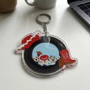 May include: A clear acrylic keychain featuring a circular design with a black tire border. The center displays a light blue sky background with three cartoon characters, a red cowboy hat, and a red cowboy boot with white stitching details.