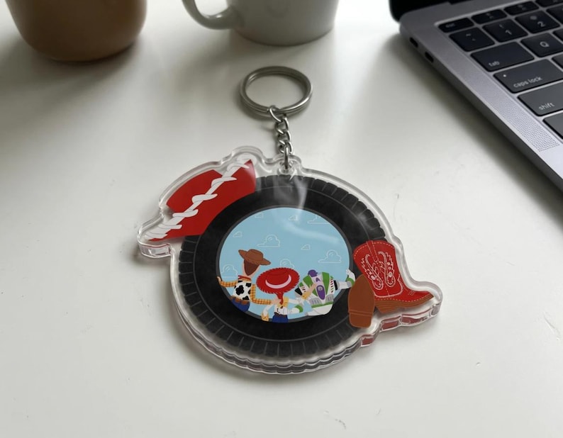 May include: A clear acrylic keychain featuring a circular design with a black tire border. The center displays a light blue sky background with three cartoon characters, a red cowboy hat, and a red cowboy boot with white stitching details.