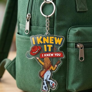 Pu&ograve; includere: Un portachiavi in acrilico trasparente con l'illustrazione di una persona dai lunghi capelli castani, che indossa una camicia a quadri gialla e pantaloni a zampa con stampa mucca, mentre tiene una chitarra. Il testo recita I KNEW IT e I KNEW YOU.