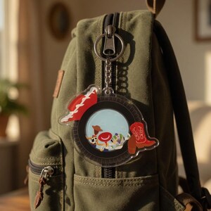 May include: A clear acrylic keychain featuring a circular design with a black tire border, a red cowboy hat, and a red cowboy boot. The center displays a cartoon illustration of characters including a cowboy and a space ranger on a light blue background.
