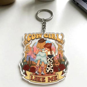 May include: A clear acrylic keychain featuring a graphic illustration of two figures in cowboy hats standing in a desert landscape with cacti and red roses. The text COW GIRL is at the top and LIKE ME is on a banner at the bottom in yellow.