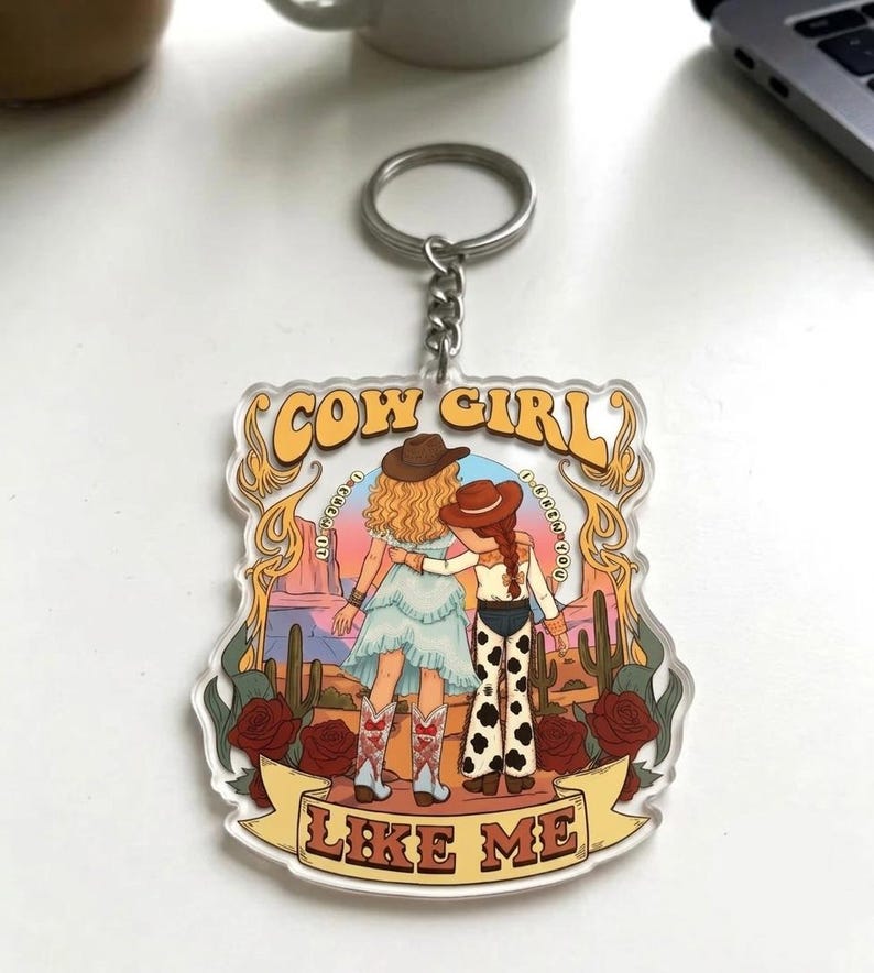 May include: A clear acrylic keychain featuring a graphic illustration of two figures in cowboy hats standing in a desert landscape with cacti and red roses. The text COW GIRL is at the top and LIKE ME is on a banner at the bottom in yellow.