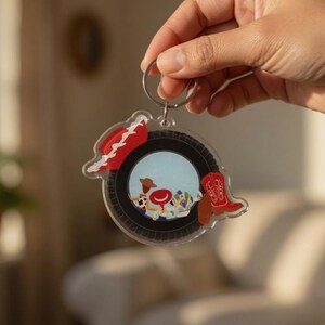 May include: A circular acrylic keychain featuring a black tire-shaped border. The center displays a colorful illustration of cartoon characters, a red cowboy hat, and a red cowboy boot. The keychain is attached to a silver metal split ring.