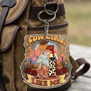 May include: A clear acrylic keychain featuring a graphic illustration of two figures in cowboy hats standing in a desert landscape with cacti and red roses. The text COW GIRL is at the top and LIKE ME is on a banner at the bottom in brown letters.