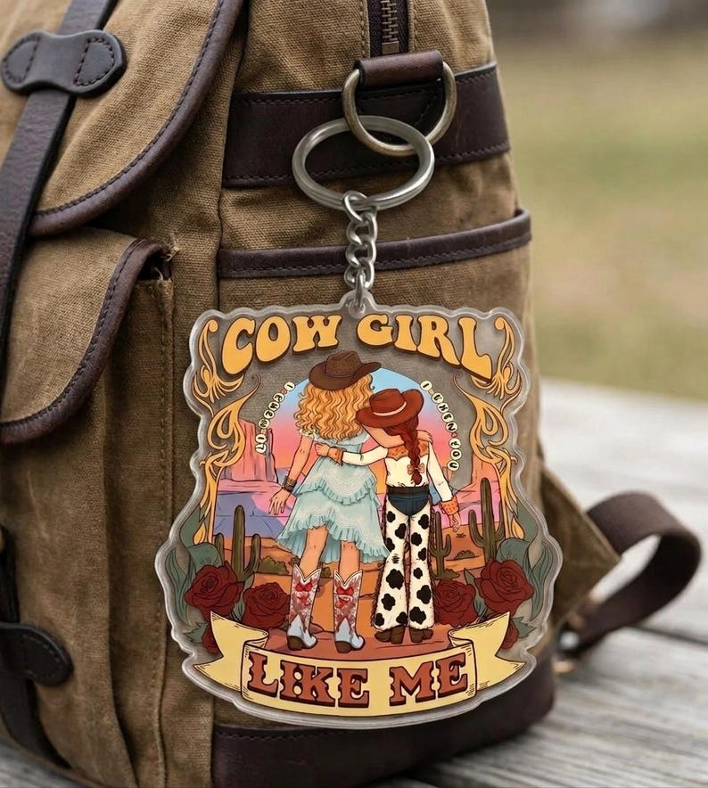 May include: A clear acrylic keychain featuring a graphic illustration of two figures in cowboy hats standing in a desert landscape with cacti and red roses. The text COW GIRL is at the top and LIKE ME is on a banner at the bottom in brown letters.