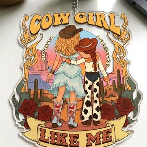 May include: A clear acrylic keychain featuring a desert illustration of two figures with arms around each other. The text reads COW GIRL at the top, I KNEW YOU I KNEW YOU on the sides, and LIKE ME at the bottom. The scene includes cacti, roses, and a sunset.