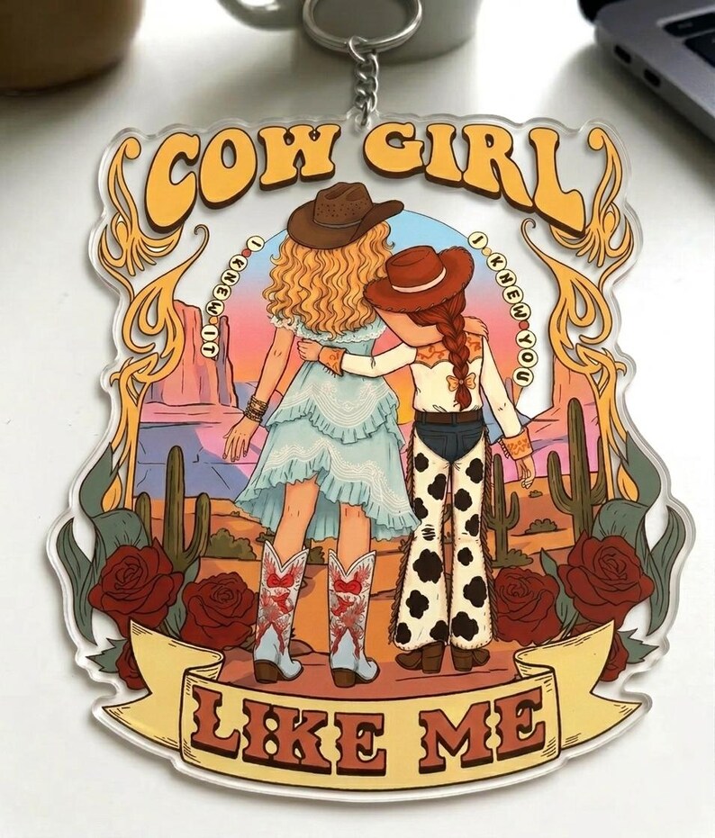 May include: A clear acrylic keychain featuring a desert illustration of two figures with arms around each other. The text reads COW GIRL at the top, I KNEW YOU I KNEW YOU on the sides, and LIKE ME at the bottom. The scene includes cacti, roses, and a sunset.