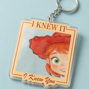 May include: A square acrylic keychain featuring a watercolor illustration of a person with red hair and a red cowboy hat. The text "I KNEW IT" is printed at the top and "I Knew You" at the bottom in a red serif font within a cream border.