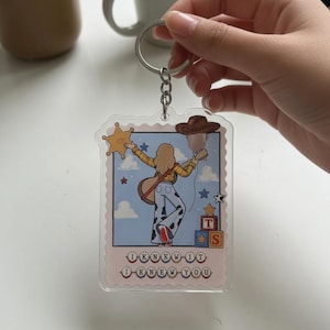 May include: A clear acrylic keychain featuring a rectangular stamp-shaped design with a cartoon illustration of a person in a yellow shirt and cow-print pants holding a guitar, a star badge, and a brown hat, with the text I KNEW IT I KNEW YOU.