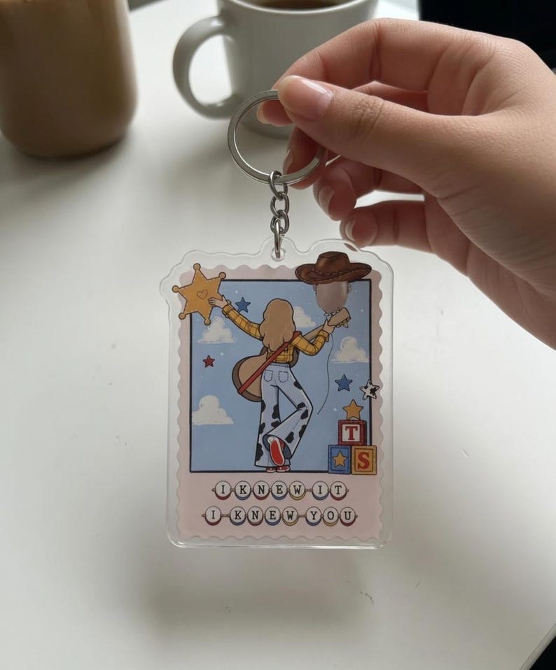 May include: A clear acrylic keychain featuring a rectangular stamp-shaped design with a cartoon illustration of a person in a yellow shirt and cow-print pants holding a guitar, a star badge, and a brown hat, with the text I KNEW IT I KNEW YOU.