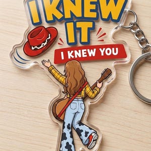 Pu&ograve; includere: Un portachiavi in acrilico trasparente con l'illustrazione di una persona dai lunghi capelli castani, camicia a quadri gialla e pantaloni a zampa con stampa mucca, che tiene una chitarra. Il testo recita I KNEW IT e I KNEW YOU, con cappello rosso.