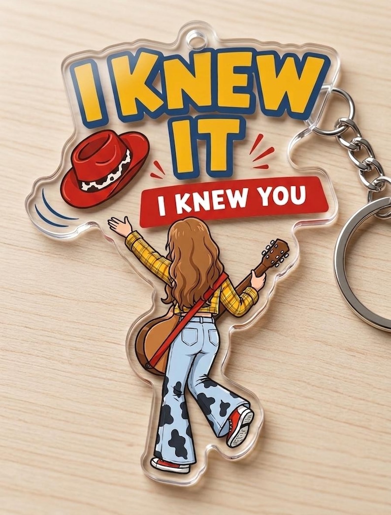 Pu&ograve; includere: Un portachiavi in acrilico trasparente con l'illustrazione di una persona dai lunghi capelli castani, camicia a quadri gialla e pantaloni a zampa con stampa mucca, che tiene una chitarra. Il testo recita I KNEW IT e I KNEW YOU, con cappello rosso.