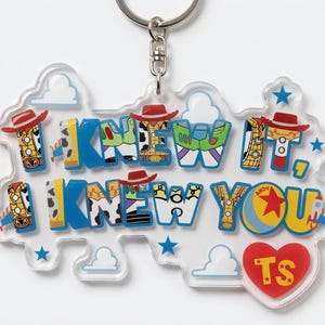 May include: A clear acrylic keyring featuring the text I KNEW IT, I KNEW YOU in blue block letters. The letters are filled with patterns of cowboy hats, space ranger suits, and sheriff badges. A red heart with the letters TS is at the bottom.