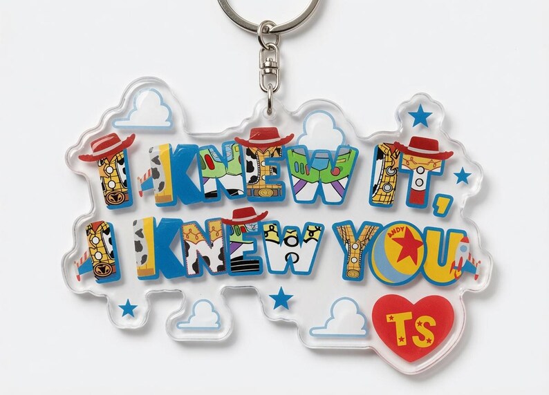 May include: A clear acrylic keyring featuring the text I KNEW IT, I KNEW YOU in blue block letters. The letters are filled with patterns of cowboy hats, space ranger suits, and sheriff badges. A red heart with the letters TS is at the bottom.