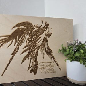 May include: A wooden panel featuring an engraved image of a Gundam-W Endless Waltz character. The artwork is detailed with wings and weaponry. The panel is light brown, with text in English and Japanese. A white and beige planter with greenery is to the right.