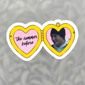 May include: A yellow heart-shaped locket sticker featuring two open halves. The left side has a pink glitter background with the black text "the summer before". The right side displays a photograph of a person wearing a black beanie and hoodie.