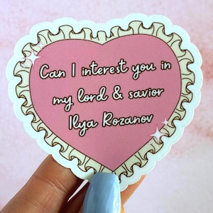 May include: A heart-shaped sticker with a ruffled cream-colored border and a pink center. The text reads: Can I interest you in my lord & savior Ilya Rozanov. Two small white four-pointed star icons are positioned on the left and right sides.