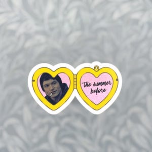 May include: A heart-shaped yellow locket sticker featuring a portrait of a person in a dark beanie with a cigarette on the left side and the text "the summer before" in black cursive script on a glittery pink background on the right side.