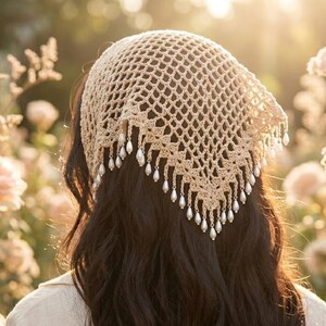 Peut inclure: Un foulard en crochet beige avec un motif triangulaire en maille ouverte et une bordure décorative ornée de petites perles blanches artificielles suspendues. Le foulard est porté sur de longs cheveux foncés, révélant la texture.