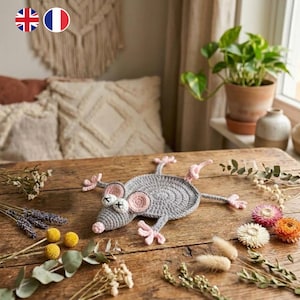 May include: A handmade gray crochet mouse coaster with pink ears, feet, and nose, and white cross-stitched eyes. The mouse is lying on a rustic wooden table, surrounded by dried flowers and greenery. A unique home decor item.