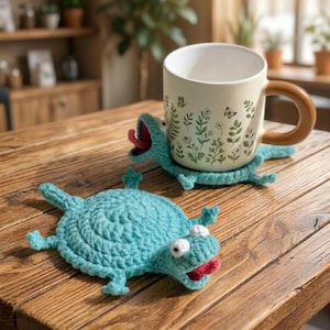 Könnte beinhalten: Eine cremefarbene Tasse mit braunem Henkel steht auf einem hellblauen, gehäkelten Eidechsen-Untersetzer. Die Tasse ist mit grünen Blumen- und Schmetterlingsmotiven verziert. Ein weiterer passender Untersetzer ist auf einem Holztisch zu sehen.