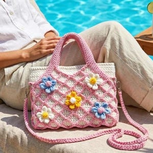 May include: A crochet handbag featuring a white base with a pink lattice overlay. The bag is decorated with five dimensional flowers in blue, yellow, and white. It includes two short top handles and a detachable long shoulder strap in pink yarn.