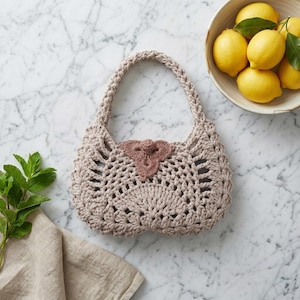 May include: A beige crocheted handbag with a braided handle and a decorative pink accent. The openwork design of the bag is visible, and it sits on a marble surface. A bowl of lemons is in the background.