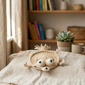 May include: A beige crocheted coaster in the shape of a whimsical creature with large, googly eyes and small arms. The coaster is round and sits on a linen surface. The background includes a bookshelf, a potted succulent, and a mug.