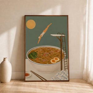 以下が含まれることがあります： ラーメンが入った大きな白い丼、麺、スープ、青菜、半分に切った卵2個が描かれた額入りアートプリント。高い茶色の飛び込み台から丼の中に飛び込む人物。黄色い太陽と赤いチリオイルが入った小さな皿が描かれています。