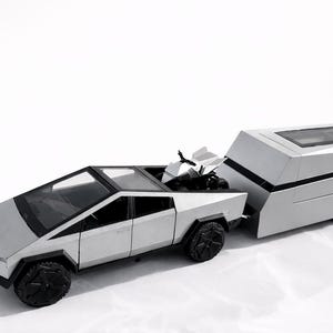 May include: A silver toy truck with a matching trailer. The truck has a unique angular design with black wheels. The trailer has a rectangular shape with a window and a solar panel on top. A small toy is in the truck bed.