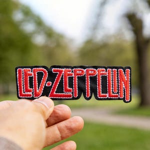 May include: A black embroidered patch with the words "LED ZEPPELIN" in red, outlined in white. The patch is rectangular and is held in front of a blurred green background.