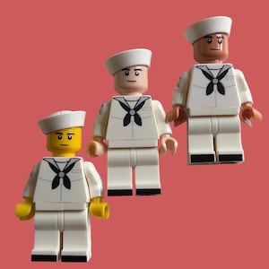 Pode incluir: Três figuras de brinquedo de plástico vestidas com uniformes de marinheiro brancos com lenços pretos e chapéus brancos. Uma figura tem a cabeça amarela, enquanto as outras duas têm tons de pele castanho claro e uma boca preta impressa.