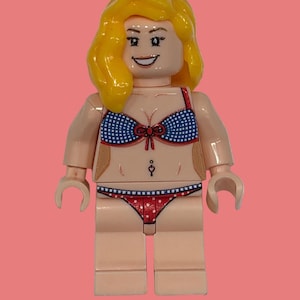 May include: A LEGO minifigure with yellow hair and a smiling expression. The figure wears a blue and white gingham bikini top with a red bow detail and matching red bikini bottoms with white polka dots, featuring a printed navel piercing.