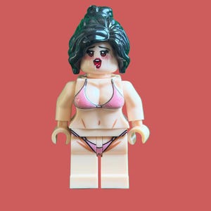 Può includere: Una minifigure in plastica con capelli verde scuro raccolti in uno chignon alto. Il personaggio ha un volto stampato con guance arrossate e bocca aperta. Indossa un top bikini rosa e uno slip coordinato con sottili laccetti stampati sul busto.
