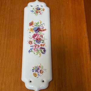 May include: A white ceramic decorative piece with a floral design. The piece has a rectangular shape with a decorative top and bottom. The floral design includes pink, blue, yellow, and purple flowers. There are two holes at the bottom for mounting.