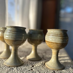 May include: A set of four ceramic goblets featuring a speckled, light beige and tan glaze. Each goblet has a rounded bowl with horizontal ridges, a slender stem, and a wide, stable circular base, showcasing a handcrafted, earthy aesthetic.
