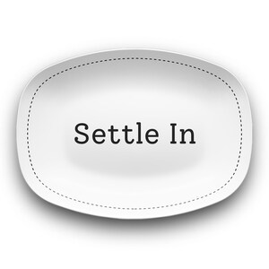 May include: A white, oval-shaped sticker with the words "Settle In" in a black, sans-serif font. The sticker has a dotted border and a slight shadow, creating a 3D effect. The background is plain white.