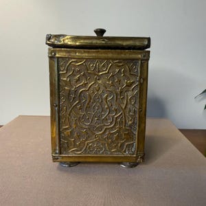 May include: A rectangular brass box with a hinged lid and a small knob. The box features intricate, embossed floral patterns on the front and sides. It rests on four small, rounded feet. The overall color is a warm, antique gold.