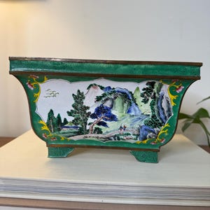 May include: Rectangular, green and white enameled planter with a landscape scene. The planter features a detailed depiction of mountains, trees, and figures. The rim and feet are green with a bronze trim. The planter is resting on a stack of books.