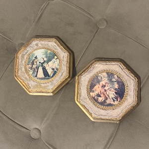 May include: Two octagonal wall plaques featuring gold-toned frames with ornate, textured borders. Each plaque contains a circular central print depicting figures in historical attire within a garden setting, framed by a decorative gold rope-like trim.