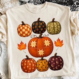 May include: A cream-colored t-shirt featuring a graphic print of seven pumpkins in various colors and patterns, including checkered, floral, vine, star, heart, polka dot, and moon designs, accompanied by two orange maple leaves on the sides.