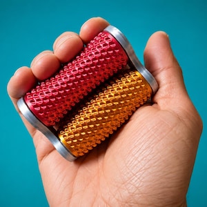 Peut inclure: Deux poignées de renforcement cylindriques, une rouge et une dorée, sont tenues dans une main sur un fond turquoise. Les poignées ont une surface texturée et des embouts argentés. Outils de fitness pour l'entraînement de la force des mains et des avant-bras.