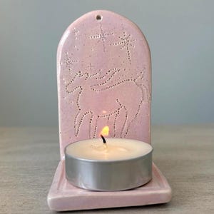 May include: A pink ceramic candle holder with a star and unicorn design. The holder has a small tray for a tea light candle. The candle is lit, casting a warm glow. The holder is a decorative item.