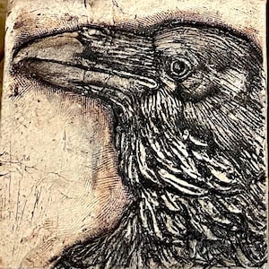May include: A black and white ceramic tile with a detailed relief sculpture of a raven's head. The raven's beak is open and its eye is visible.