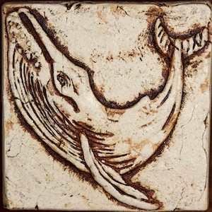 May include: A white ceramic tile with a brown relief image of a whale jumping out of the water. The whale is facing to the right and has a detailed design.