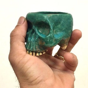 May include: A green ceramic skull planter with white teeth. The planter is in the shape of a human skull and has a small opening at the top for planting.